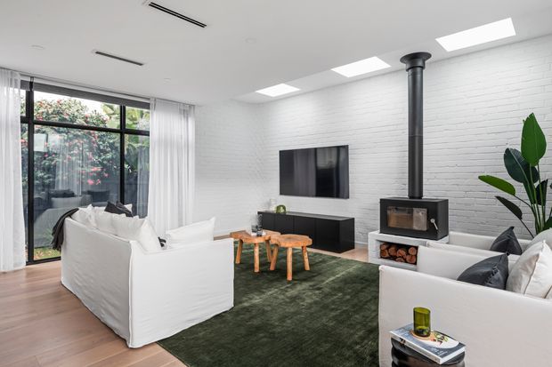 Bright contemporary living room with white-painted brick walls, exposed black wood-burning stove and flue, large black-framed glazed doors, white slipcovered sofas, a dark green rug, and timber flooring. A wall-mounted television and low black media cabinet face the seating area, while indoor plants and garden views add a natural connection to the outdoors.