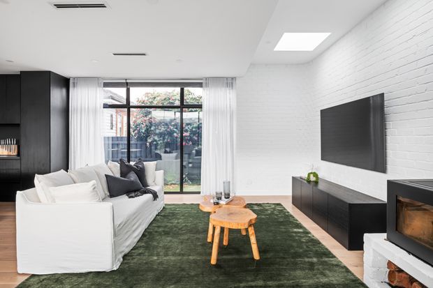 Bright contemporary living room with a white slipcovered sofa, dark built-in cabinetry, a large wall-mounted television, and a black fireplace set against a white-painted brick feature wall. A green area rug and timber coffee tables add warmth, while full-height glazing and sheer curtains connect the room to a small outdoor area. A skylight and recessed ceiling lighting enhance the airy space.