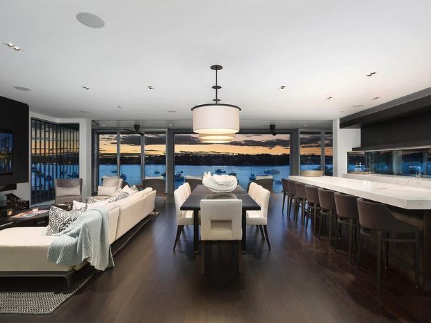 Open-plan waterfront interior combining a living room, dining area and kitchen. The room features dark timber flooring, white upholstered seating, a long stone kitchen island with bar stools, extensive floor-to-ceiling glazing, and a large central pendant light. Water and a sunset are visible beyond the windows.
