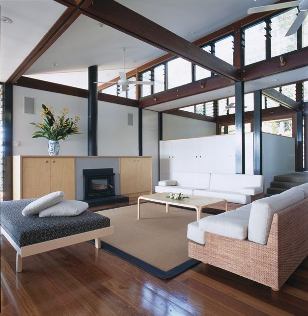 A spacious, light-filled living room with a dramatic double-height ceiling and exposed dark timber post-and-beam structure. The room features polished dark timber flooring, white walls, high clerestory windows, a black cylindrical fireplace flue, and a low timber fireplace cabinet with a built-in hearth. White sofas, a woven rattan armchair, a patterned chaise, and a slim light-coloured coffee table create a relaxed contemporary interior.