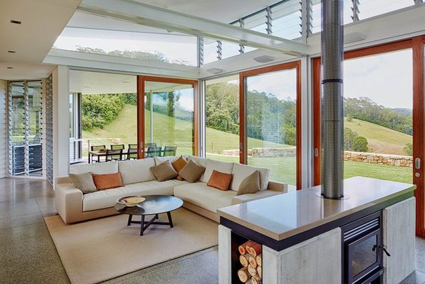 Bright, open-plan living room with a large sectional sofa, coffee tables and full-height glazing overlooking a green rural landscape. The room features a double-height glazed corner, exposed white structural beams, timber-framed windows and doors, a modern fireplace with a cylindrical flue, and a kitchen or joinery island in the foreground. The interior is airy and contemporary with a neutral palette and warm timber accents.