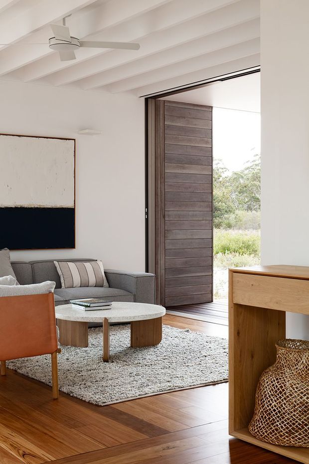 A bright, warm living room with white walls and exposed painted ceiling beams. The room features a grey sofa, leather accent chair, round light-stone coffee table, textured rug, timber flooring, and a substantial timber-framed open doorway with a dark horizontal-board timber door leading outdoors. A timber console or built-in cabinet occupies the right foreground, with woven storage baskets below. The space has a relaxed contemporary coastal character with strong natural materials and indoor-outdoor connection.