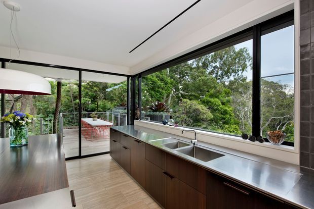 A contemporary kitchen with a long dark timber island and matching peninsula cabinetry, stainless-steel benchtops, an integrated double sink, and a tiled feature wall. Full-height black-framed glazing and sliding doors open the space to a leafy outdoor terrace, while a large white pendant light hangs over the island. The room is bright, open, and strongly connected to the garden.