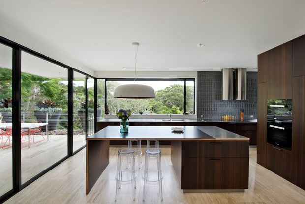 Contemporary open-plan kitchen with a large dark timber island, integrated appliances, grey tiled splashback, pale timber flooring, and extensive black-framed glazing opening toward a leafy outdoor area. A sculptural pendant hangs above the island, with transparent bar stools beneath the seating overhang.