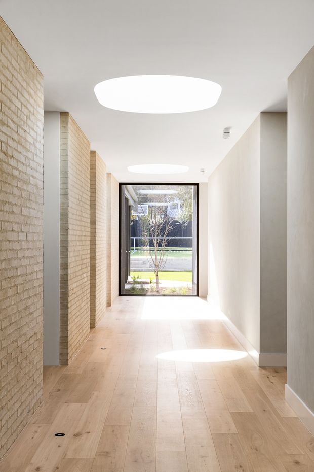 Bright minimalist hallway with pale timber plank flooring, exposed light brick walls, smooth white plaster walls and ceiling, and large oval skylights casting pools of sunlight. A full-height glazed door at the end opens toward a landscaped outdoor area, while recessed openings and simple detailing create a clean, linear passage.