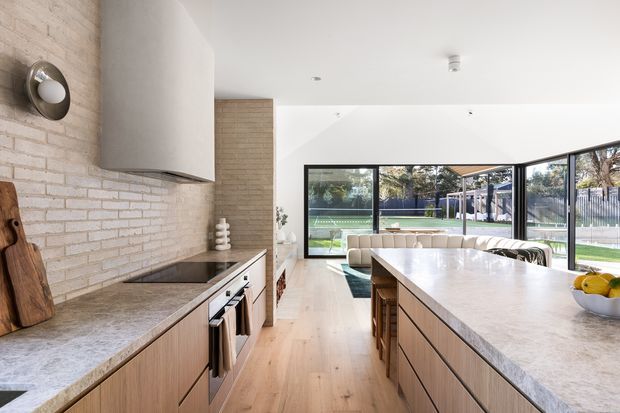 Bright open-plan kitchen and living area with a large central island, pale timber cabinetry, and a light stone benchtop. The kitchen features a white-painted brick splashback wall, integrated cabinetry, stainless-steel appliances, and timber flooring. Floor-to-ceiling black-framed glazing opens the room visually to a landscaped outdoor area and pool, while a curved upholstered sofa sits beyond the island.