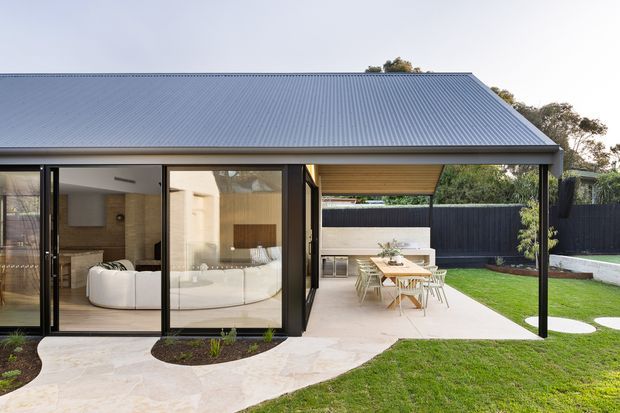 Contemporary single-storey home viewed from the landscaped backyard, featuring a large covered outdoor entertaining area with a pitched corrugated metal roof, slender dark framing, extensive sliding glass doors, and warm timber soffit lining. The open plan interior shows a white sectional sofa and timber-toned finishes, while the exterior includes a paved terrace, outdoor dining setting, lawn, garden beds, and a compact built-in outdoor kitchen or barbecue zone.