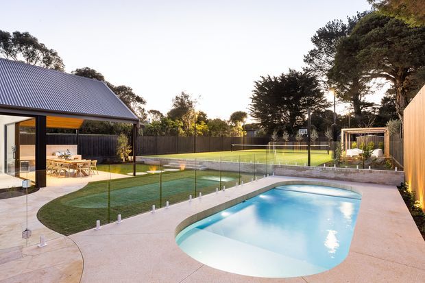 A landscaped residential backyard viewed from an elevated patio or terrace, featuring a curved swimming pool, a second fenced sports court or recreational area, lawn, paved surrounds, and a modern pavilion-style outdoor dining space. The property includes dark vertical fencing, timber screening, extensive mature trees, and a detached structure with a steep corrugated metal gable roof and glazed walls.