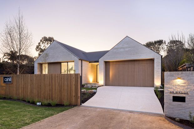 Street-facing photograph of a contemporary single-storey residence with intersecting pitched roof forms, pale masonry walls, warm timber garage and entry doors, and a broad concrete driveway. A timber privacy fence, landscaped front garden, and illuminated masonry letterbox frame the frontage at dusk.