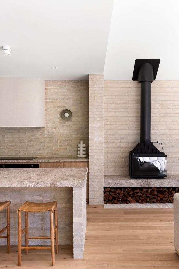A warm contemporary open-plan interior combining a compact kitchen and living area. Pale exposed brickwork forms the walls and a central dividing pier, with a stone-look kitchen benchtop, light timber cabinetry and flooring, and a prominent black wood-burning stove set into a built-in hearth with stacked firewood storage below. A timber bar stool and recessed ceiling lighting complete the scene.