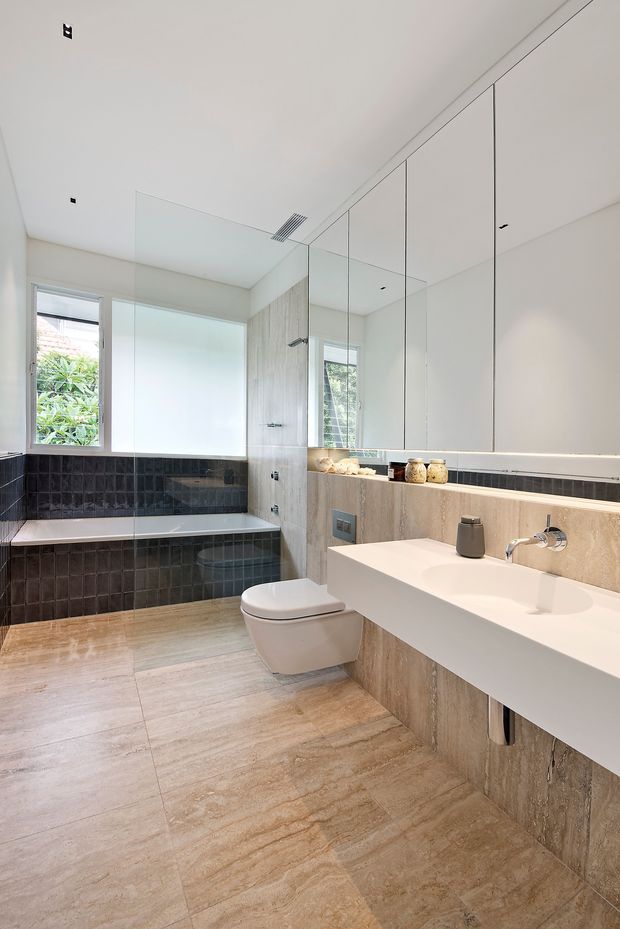 Bright contemporary bathroom featuring a wall-hung toilet, large floating double vanity with integrated basins, full-height mirrored cabinetry, a freestanding bathtub set within dark textured wall tiles, and a frameless glass shower enclosure. The room uses warm beige stone-look floor and wall finishes with crisp white surfaces and concealed ceiling lighting.