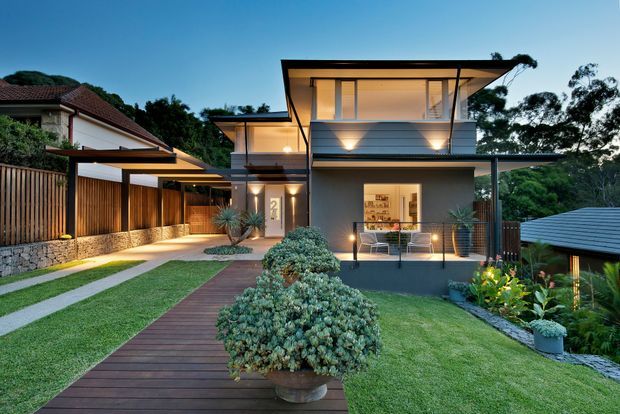 Twilight exterior view of a contemporary multi-level residence with broad overhanging flat roof planes, large glazed openings, a covered upper balcony, and warm architectural lighting. A timber boardwalk leads through a landscaped front garden toward the entry and elevated living spaces. Vertical timber screening, stone retaining walls, clipped shrubs, and outdoor planting frame the house; neighbouring rooflines and dense trees are visible in the background.