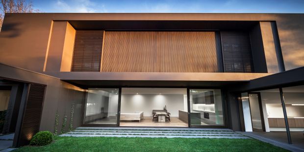 A contemporary two-storey residence viewed from the landscaped front garden at dusk. The dark, rectilinear facade has broad projecting roof and wall planes, extensive black-framed glazing, and a prominent upper-level timber slatted screen. The ground floor opens toward a paved terrace and reveals a sparsely furnished living or dining area, while recessed exterior lighting creates a warm, refined atmosphere.