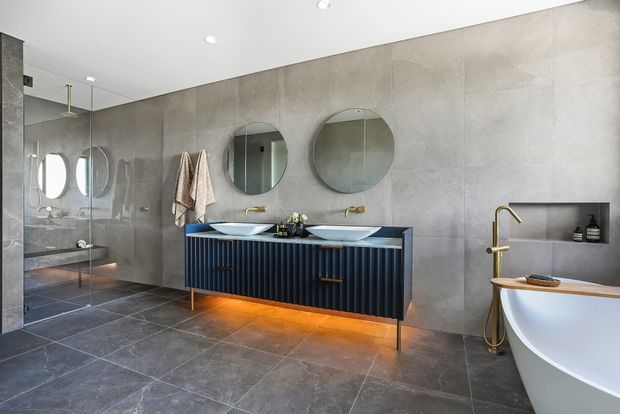 Contemporary bathroom with large-format grey concrete-look wall and floor tiles, a floating navy fluted double vanity with two basins, round mirrors, brass tapware, a glass-enclosed shower, and a freestanding white bathtub. Warm under-vanity lighting adds contrast to the cool grey finishes.
