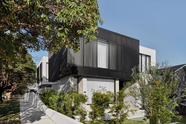 Contemporary multi-level residential house viewed from the street, featuring crisp white rendered walls, a prominent dark vertical slatted screen enclosing the upper level, large glazed openings, and landscaped garden beds along a curved pale boundary wall and sidewalk. Mature trees partially frame the facade.
