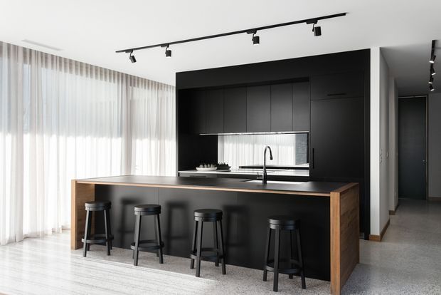 Contemporary open-plan kitchen dominated by a large black island with timber end panels and four black stools. A full-height black cabinetry wall incorporates a sink, dark benchtop and mirrored splashback, while sheer floor-to-ceiling curtains provide abundant natural light. Track lighting and polished light flooring create a clean, refined monochrome interior.