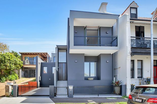 Street-level exterior view of a narrow modern two-storey townhouse with a grey rendered facade, projecting upper balcony, black metal fencing and balcony balustrades, and large glazed openings. Traditional attached neighbouring homes are visible on either side.