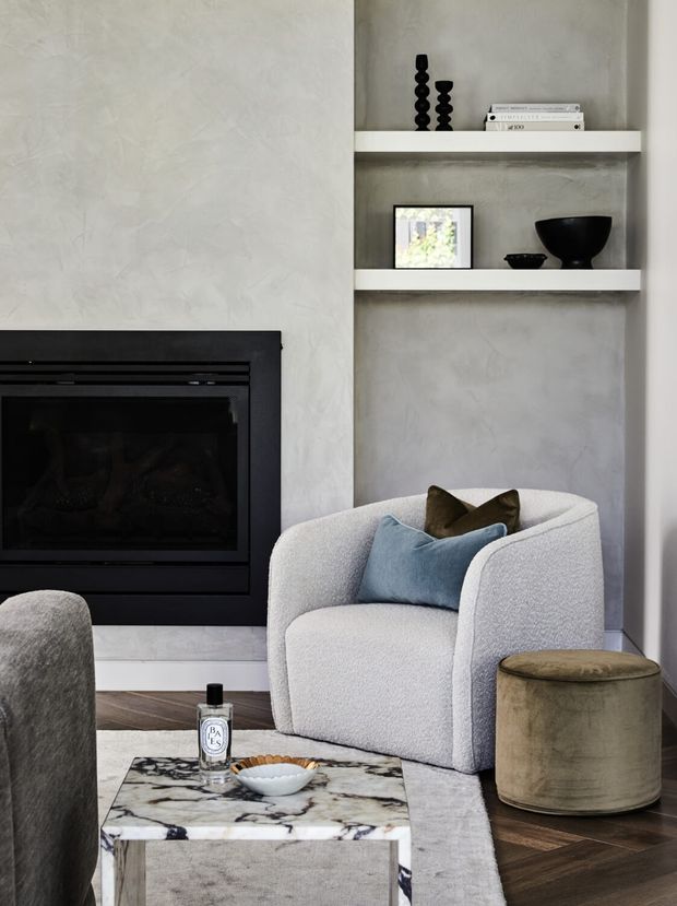 A refined contemporary living room vignette featuring a black built-in fireplace, textured pale plaster walls, recessed open shelving with minimalist black ceramics and books, a curved upholstered armchair, a marble coffee table, and dark herringbone timber flooring. The palette is neutral with soft grey, white, black, and muted olive accents.