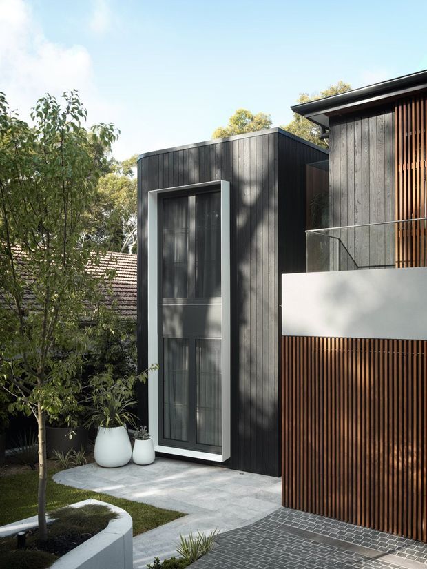 A contemporary residential exterior viewed from a landscaped front approach. The facade combines dark vertical timber cladding, a tall framed glazed opening, warm timber screening, a white rendered balcony parapet, and a low-pitched flat roof. Grey paving, curved raised planters, mature planting, and a brick-paved driveway complete the scene.
