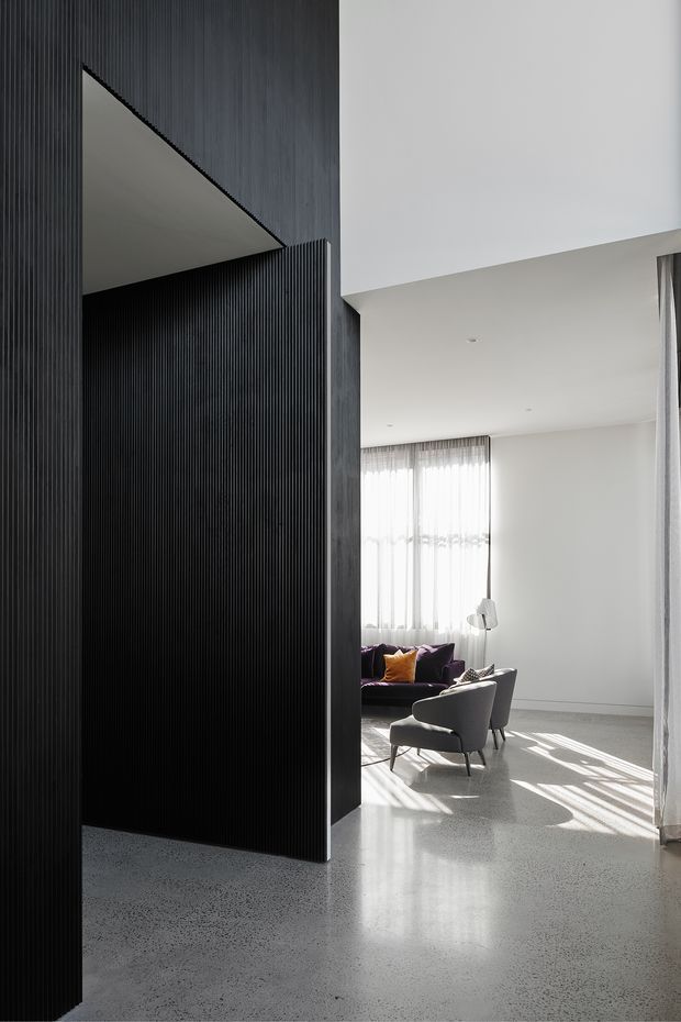 A dramatic contemporary interior viewed from a dark, vertically ribbed architectural partition into a bright living room. The living area has white walls and ceiling, polished speckled concrete flooring, full-height sheer curtains, a dark sofa with a mustard cushion, two grey lounge chairs, and strong sunlight casting linear shadows across the floor. The space features a high ceiling and a strong contrast between the charcoal partition and the light-filled room.