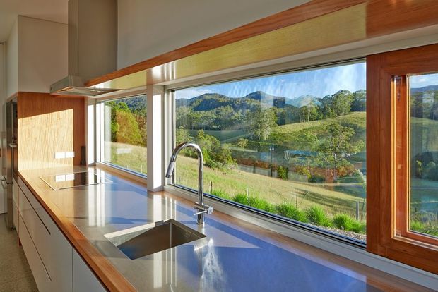 A bright contemporary kitchen viewed along a long benchtop, featuring extensive timber-framed glazing overlooking a green rural landscape and water. The kitchen includes sleek pale cabinetry, a polished grey-blue benchtop, integrated cooktop, undermount stainless-steel sink, and a curved chrome mixer tap. Warm timber trims and window frames contrast with the light interior.