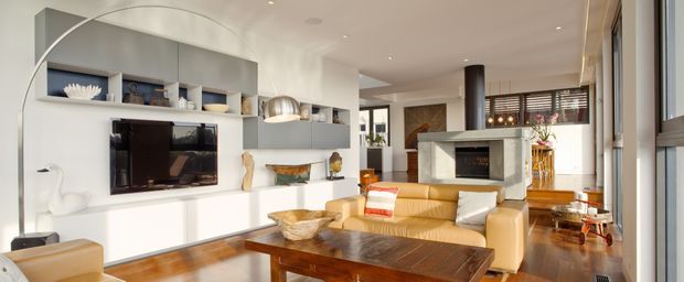 Bright contemporary living room viewed from near a glazed opening, featuring a built-in white entertainment and display wall, flat-screen television, tan leather sofa, dark timber coffee table, timber flooring, recessed ceiling lights, and a prominent curved arc floor lamp. A stone-clad fireplace and dining area are visible deeper in the open-plan space.