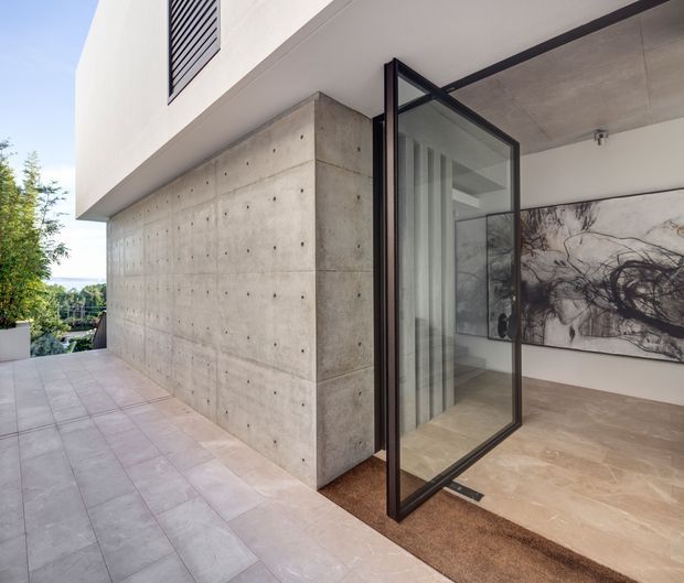 A contemporary indoor-outdoor threshold with a large pivoting black-framed glazed door opening from a tiled terrace into a minimalist interior. The exterior wall is exposed board-formed concrete with visible tie holes, while the interior features a smooth concrete ceiling, pale stone or tile flooring, white walls, and an abstract monochrome artwork. A landscaped outlook is visible beyond the terrace.