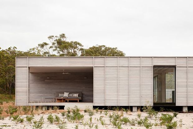 Single-storey contemporary rectangular dwelling set in a sparse natural landscape. The facade is clad in pale horizontal weatherboard-like panels with strong linear battens, a large recessed covered outdoor living area, and a tall glazed entry opening. The sheltered recess has dark timber wall and ceiling finishes, a ceiling fan, and built-in seating. Low coastal vegetation and sandy ground surround the building.