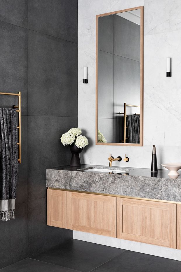 A refined contemporary bathroom vanity featuring a floating light timber cabinet, a thick mottled grey stone benchtop with integrated basin, brass wall-mounted tapware and towel rail, large black and white marble-look wall tiles, and a timber-framed rectangular mirror. Black floor and wall tiles create a strong contrast, complemented by minimalist cylindrical wall lights and restrained decorative accessories.