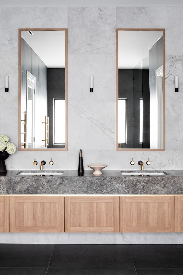 A refined contemporary bathroom vanity with two integrated basins, wall-mounted brass and black tapware, large rectangular timber-framed mirrors, pale marble-look wall panels, a dark grey stone benchtop, and warm timber cabinetry. Black floor tiles and minimalist cylindrical wall sconces create a high-contrast, sophisticated scheme.