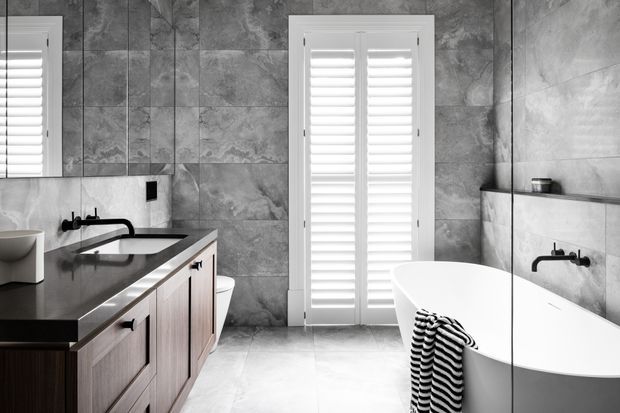 A refined contemporary bathroom featuring a freestanding white bathtub, dark stone vanity benchtop with timber cabinetry, wall-mounted black tapware, large grey stone-look wall tiles, and white plantation shutters. A large mirror reflects the vanity and bath area, while a striped towel adds a small graphic accent.