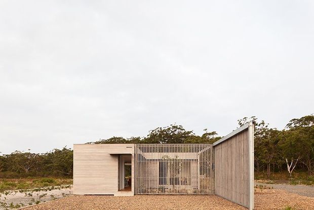A low, single-storey modern pavilion set in a sparse coastal or bush landscape. The structure has pale horizontal timber-clad walls, a tall screened courtyard or entry made from closely spaced vertical slats, and a simple pitched or angled roof form. The foreground is covered with pale gravel and sand, with native vegetation beyond under an overcast sky.