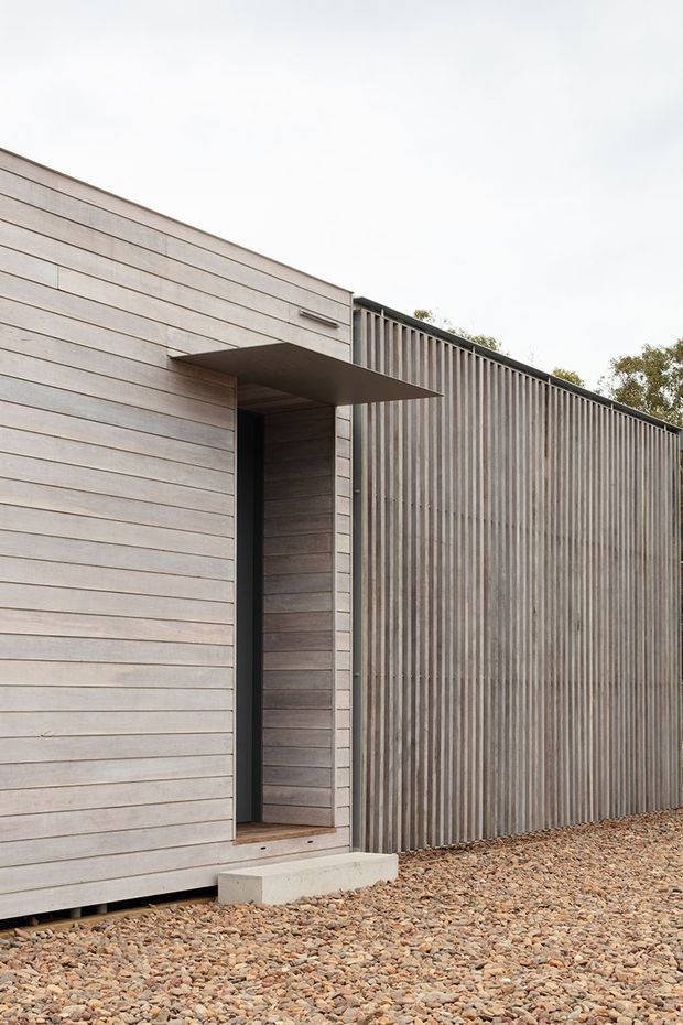 Minimal modern exterior detail showing a narrow recessed entry door beneath a projecting flat canopy. The facade is clad in horizontal pale timber boards, contrasted with a tall vertically ribbed wall or screen, and set above a gravel-covered ground plane.