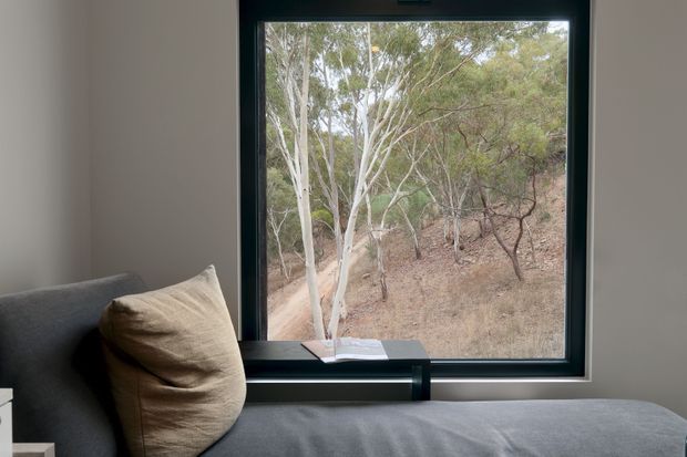 A quiet, softly lit interior corner with a grey upholstered daybed or sofa beside a large black-framed window. A beige cushion rests against the seating, while the window overlooks a dry eucalyptus woodland landscape. The room has a restrained, contemporary feel with neutral finishes and a strong connection to nature.