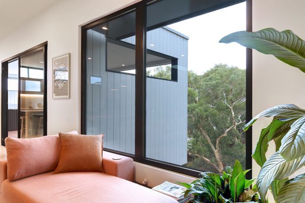 A bright contemporary living room with a large peach-toned sofa positioned beside expansive dark-framed windows. The glazing overlooks a landscaped leafy setting and a modern vertically clad building volume, while indoor tropical plants, framed artwork, and a low window ledge add warmth and texture.