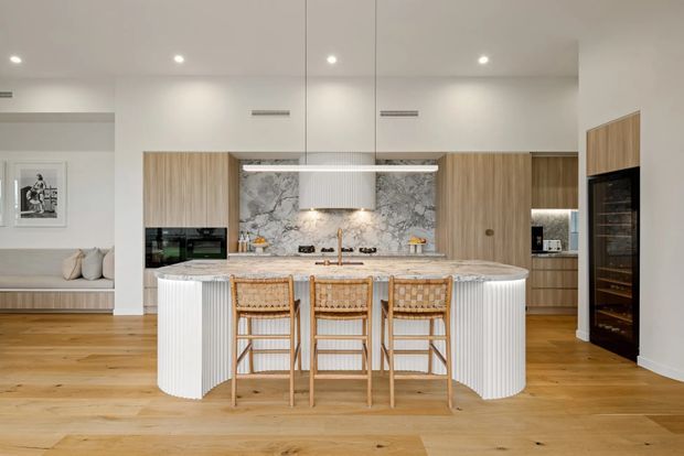 Bright contemporary kitchen with a large white fluted island, three woven timber bar stools, warm timber flooring, integrated timber cabinetry, marble-look stone splashback and benchtops, and a concealed rangehood. A built-in seating nook and framed artwork are visible to the left, while tall integrated appliances and additional cabinetry occupy the right side.