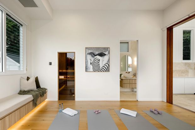 Bright contemporary open room with pale timber flooring, a built-in window seat, exposed white walls, and several yoga mats arranged on the floor. A compact sauna or timber-lined room is visible through one doorway, while another doorway opens to a bathroom with a vanity. The space is minimal and wellness-oriented, with white finishes, warm wood, and abundant natural light.