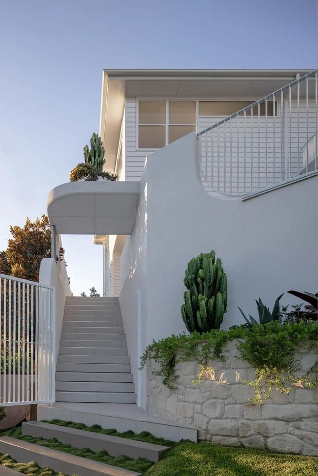 A bright exterior view of a contemporary multi-level residence, focused on a stairway leading to an elevated entry and upper terrace. The white rendered facade features horizontal cladding, slim metal-framed glazing, white grid-pattern balustrades, and a curved projecting canopy. Layered landscaping includes sculptural cacti, succulents, low groundcover, and a pale natural-stone retaining wall.