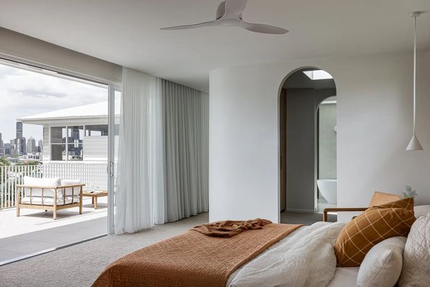 Bright contemporary bedroom with a large glazed opening to a balcony or terrace, sheer full-height curtains, a low-profile ceiling fan, and a rounded-arch doorway leading toward a bathroom area. The room has a neutral white and grey base with warm brown bedding and cushions, and a city view beyond the outdoor railing.