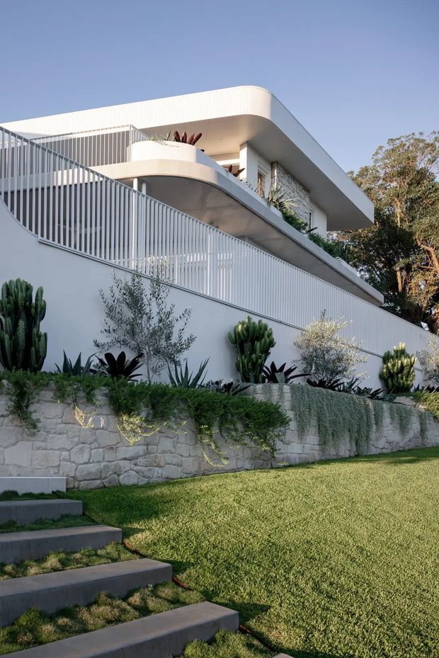 A contemporary multi-level white residence viewed from the landscaped garden below. The building features rounded roof and balcony edges, broad overhanging eaves, extensive white vertical balustrade screening, and layered terraces. A pale natural-stone retaining wall supports a planted garden with sculptural succulents and cascading greenery, while dark concrete stepping stairs cross the lawn in the foreground.