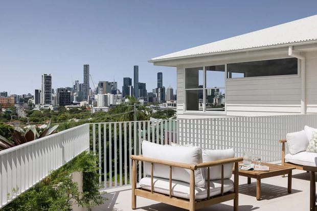 A bright elevated outdoor terrace overlooking a dense city skyline. The terrace features white vertical balustrades, pale decking, a light-coloured contemporary building enclosure with broad glazing and horizontal cladding, and timber-framed outdoor lounge furniture with neutral cushions and a low coffee table.