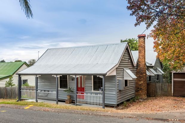 Street-facing view of a compact single-storey cottage with a corrugated metal gable roof, grey horizontal weatherboard cladding, a covered front verandah, white railings and trim, and a distinctive red front door. A tall brick chimney rises beside the roof, with neighbouring cottages and mature trees visible around it.