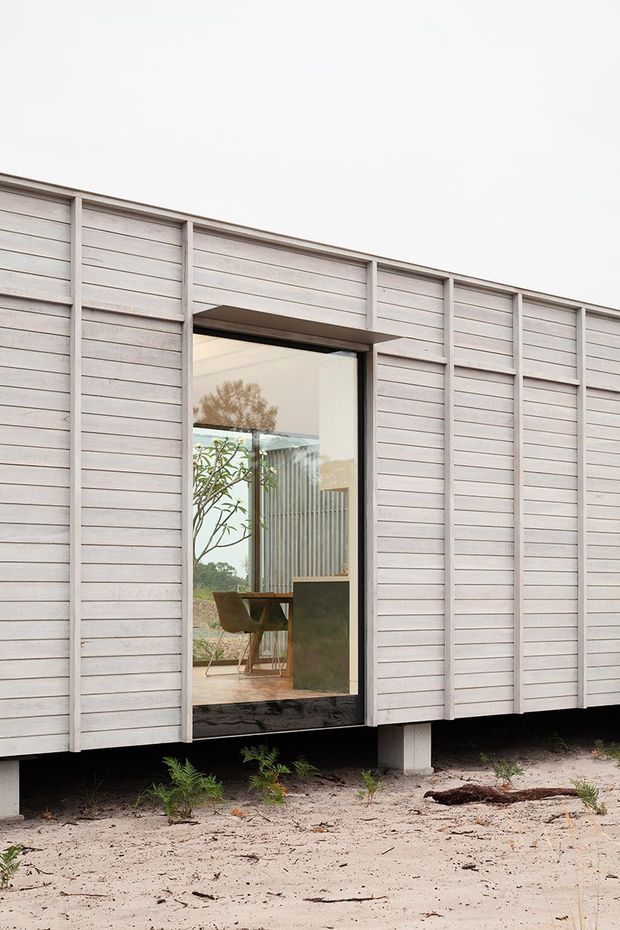 Exterior view of a compact elevated building clad in pale horizontal weatherboards, with a large glazed opening revealing a simply furnished interior. The structure is raised above sandy ground on narrow supports, and the facade has a clean, rectilinear composition with vertical framing battens.