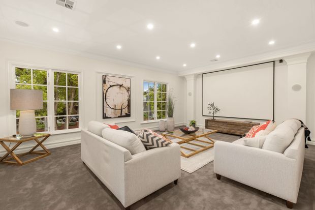 Bright, spacious formal living room with white upholstered sofas and armchairs arranged around a layered glass-and-metal coffee table. The room features grey carpet, white walls and ceiling with recessed lighting, tall multi-pane windows, classic wall moulding, a large wall-mounted projection screen, a timber media console, indoor plants, and neutral contemporary decor with red and black accents.