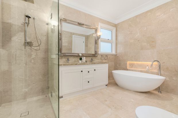 A spacious, refined bathroom featuring full-height beige stone-look wall and floor tiles, a frameless glass walk-in shower, white built-in vanity cabinetry with a stone benchtop, a large ornate-framed mirror, a freestanding oval bathtub, and chrome fixtures. Warm concealed lighting highlights the bath wall niche, while a window provides natural light.
