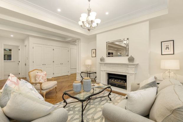 Elegant, light-filled formal living room with cream upholstered sofas and armchairs arranged around a glass-top coffee table. The room features a white carved fireplace mantel, built-in white cabinetry and wardrobes, herringbone timber flooring, layered rugs, a crystal chandelier, table lamps, wall art, and a mirrored overmantel. The overall presentation is refined and traditional with soft neutral furnishings.
