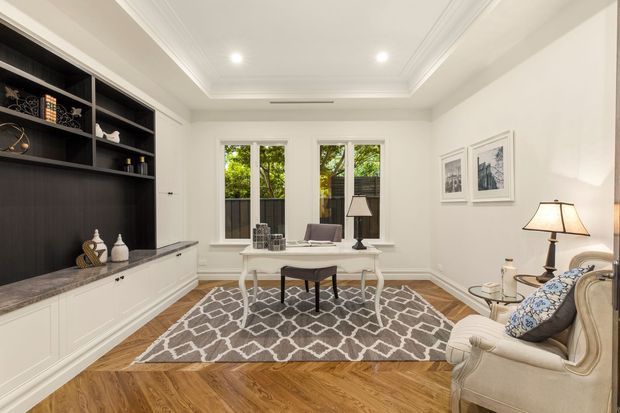 Elegant traditional sitting room with a built-in dark display and media wall, white lower cabinetry, marble-look benchtop, ornate ceiling cornices, timber parquet flooring, and a patterned area rug. A small writing desk sits in front of three windows, with upholstered armchairs, table lamps, framed artwork, and classic decorative furnishings.