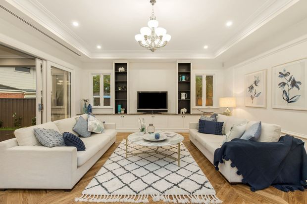 Bright, refined living room with white sofas, navy and pale blue cushions, a patterned area rug, pale timber flooring, built-in white media cabinetry, framed botanical artwork, and a central crystal chandelier. Glazed doors open to an exterior area, while recessed ceiling lights and decorative cornices create a polished, traditional coastal feel.