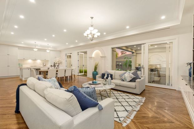 Bright, spacious open-plan living and dining area with a white kitchen at the rear. The room features white upholstered sofas, a pale neutral palette, herringbone timber flooring, recessed ceiling lights, decorative wall mouldings, a chandelier, and large glazed doors opening toward an outdoor courtyard or garden. A patterned rug and blue cushions provide subtle contrast.