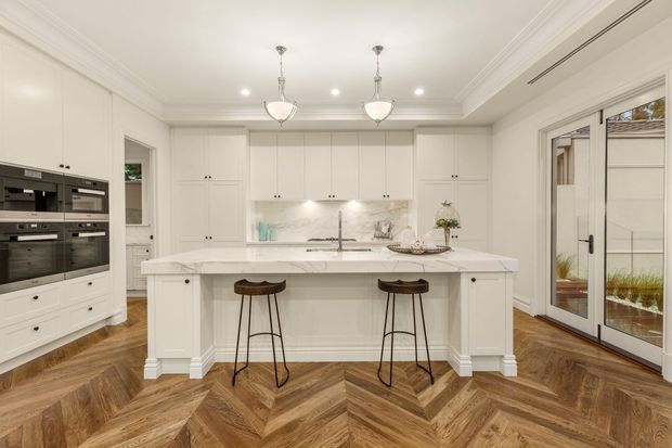 A bright, refined traditional kitchen with extensive white shaker-style cabinetry, integrated double ovens, a large central island with a pale stone benchtop and two bar stools, and a herringbone timber floor. Decorative pendant lights, ceiling cornices, and wall panel detailing add a classic character, while glazed doors open to an exterior area.